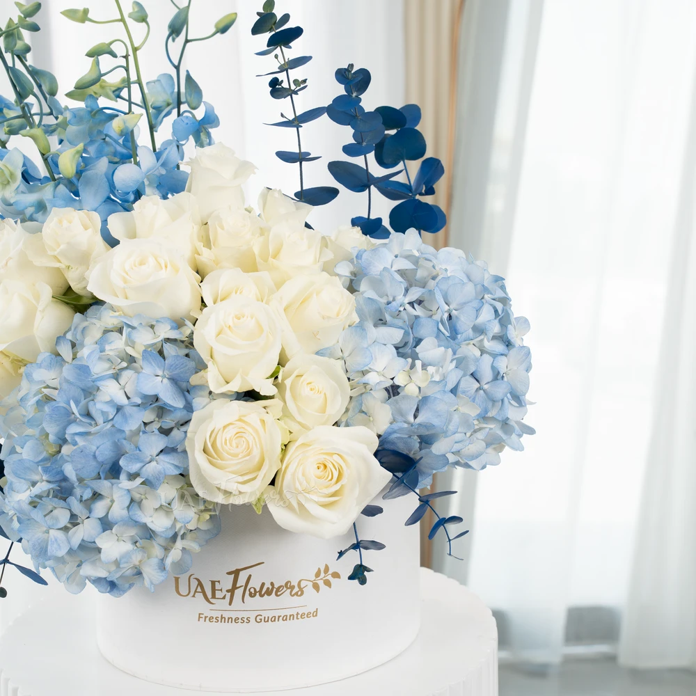 A box arrangement of 2 blue hydrangeas, 20 white roses, 5 blue orchids and blue sprayed eucalyptus leaf.
