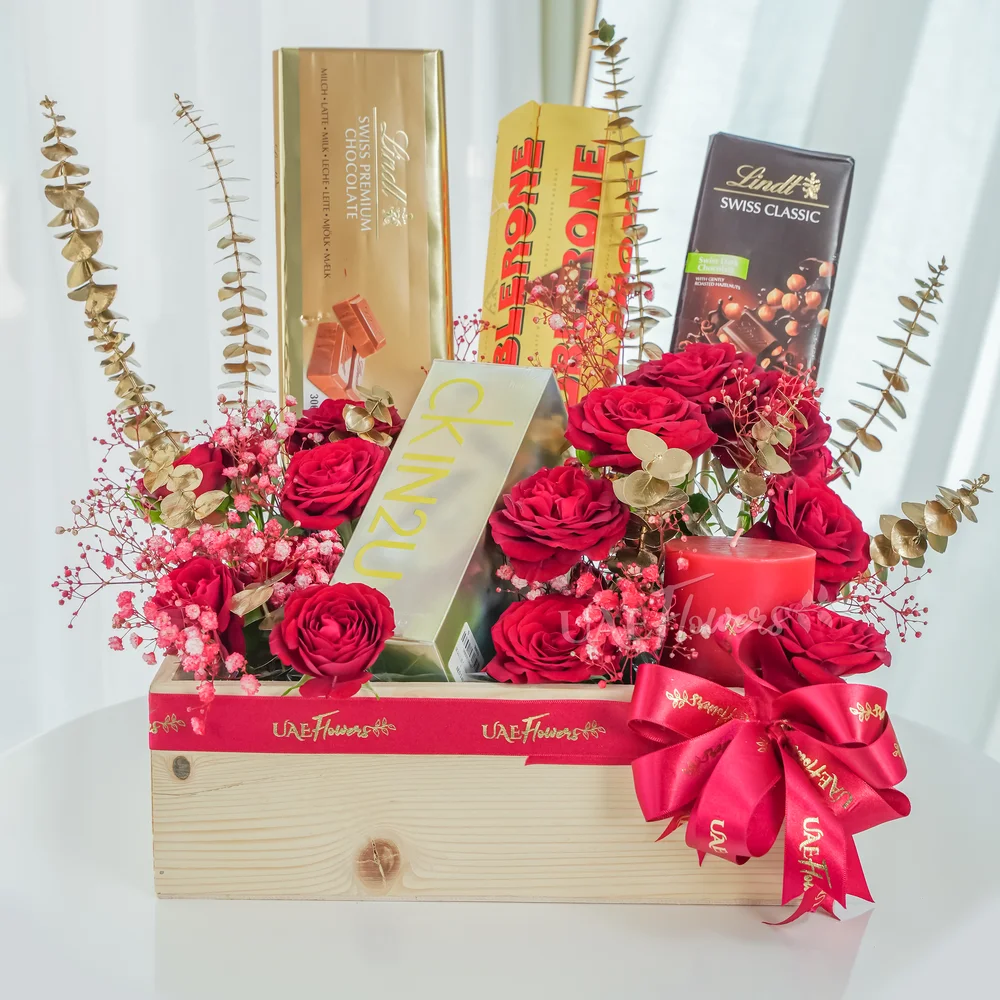 A wooden box arrangement of 12 red roses, 6 Pcs Toblerone Chocolates, Lindt Swiss Dark Chocolate Classic 100g, Lindt Swiss Premium Chocolate Bar 300g and CK In2u L EDT 100ML perfume with gold sprayed eucalyptus leaf and baby's breath.