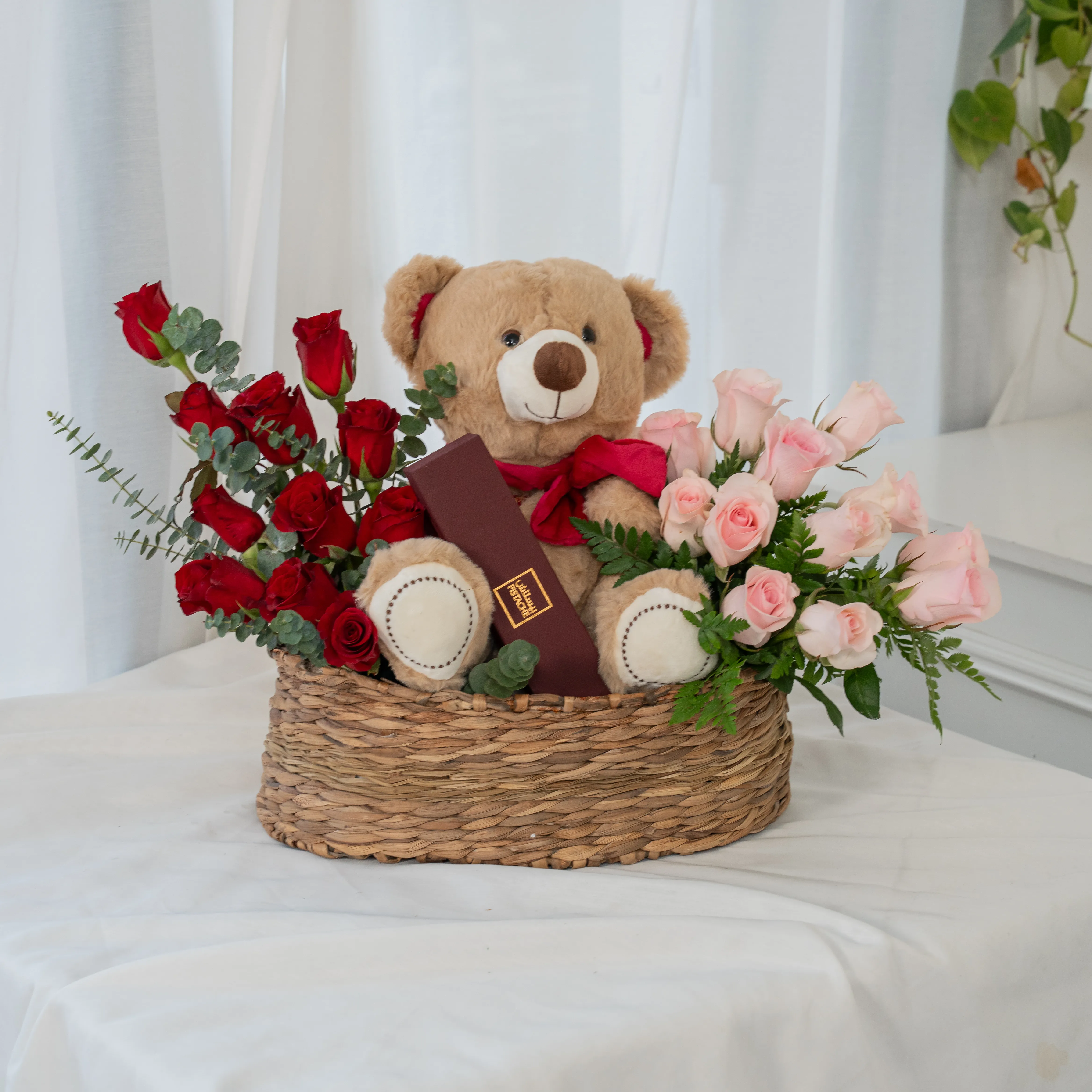 Basket arrangement of 11 red roses, 11 pink roses, small brown teddy bear and 6 Pcs Pistache Petite Indulgence Chocolate Box with green fillers.