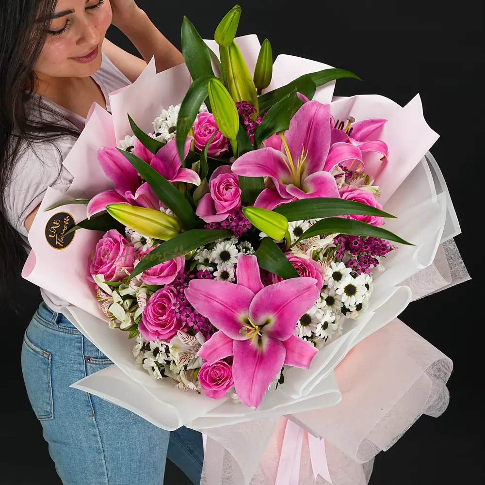 A bouquet of 3 pink lilies, 10 pink roses, 2 white alstromeria, 2 white chrysanthemums and wax flowers in pink wrapping paper tied with pink ribbon